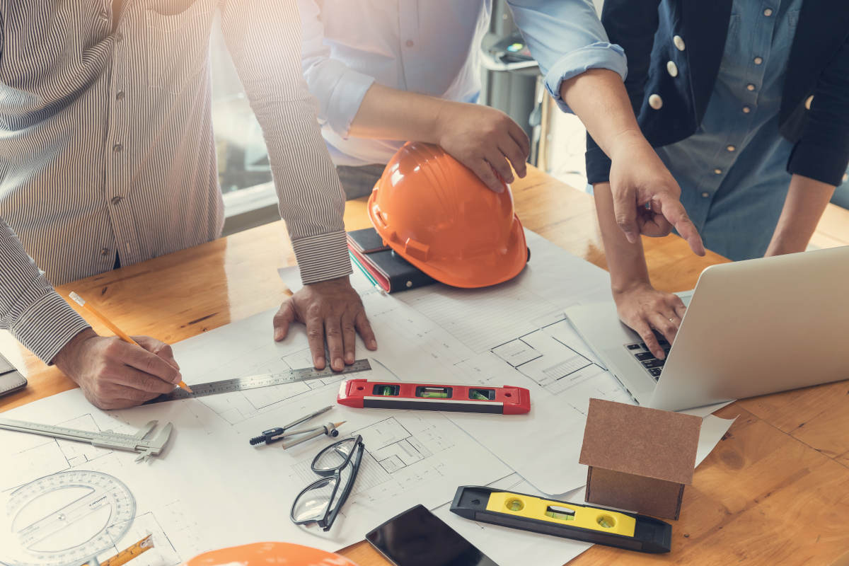 Team discussing blueprints with computer, hard hat, rules, glasses spread out on table